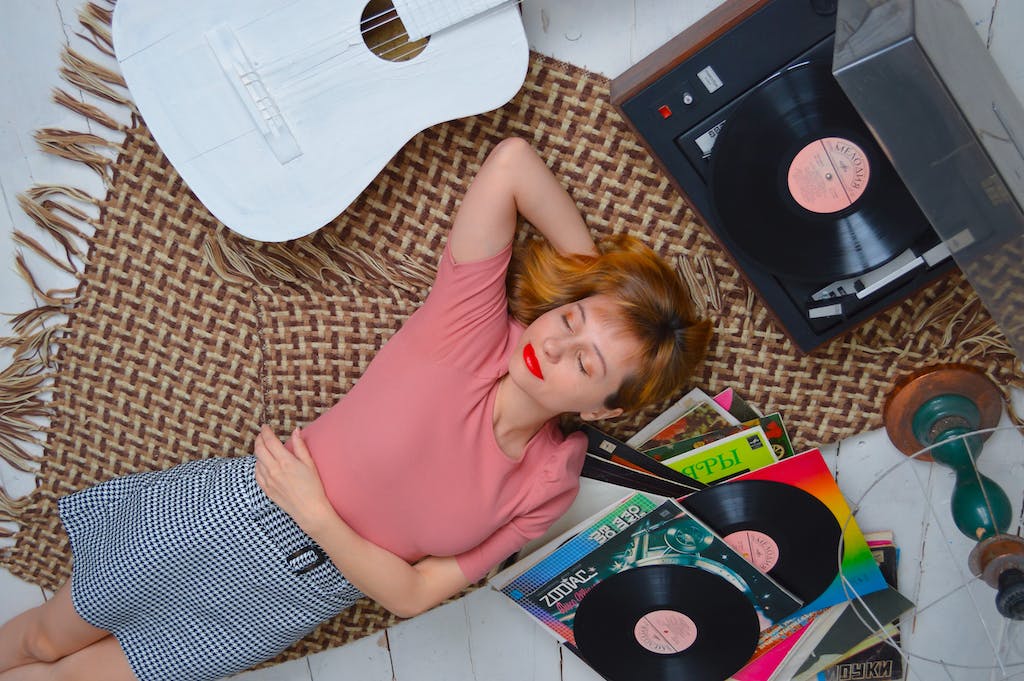 Top view dreamy young female with red lips wearing casual wear lying on floor with eyes closed and hand behind head and listening to vinyl record player